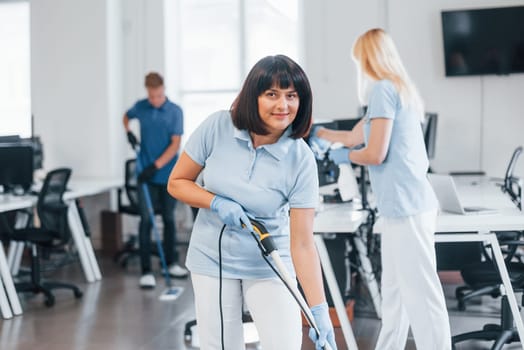 Woman uses vacuum cleaner. Group of workers clean modern office together at daytime.