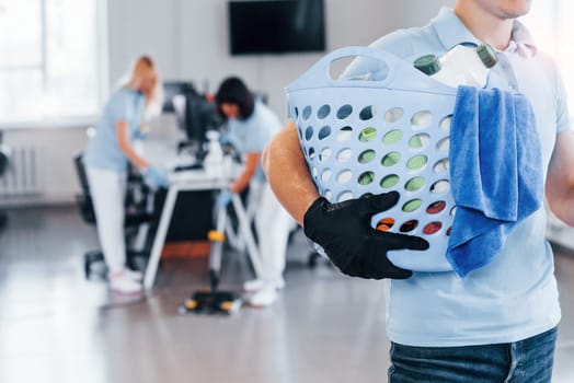 Man holds basket. Group of workers clean modern office together at daytime.