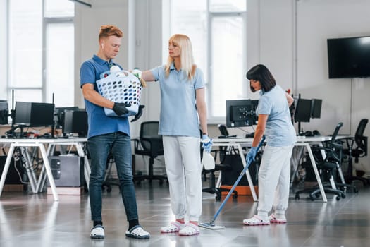 Man holds basket. Group of workers clean modern office together at daytime.
