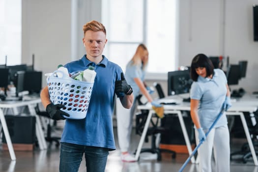 Man holds basket. Group of workers clean modern office together at daytime.