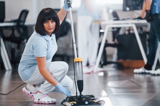 Woman uses vacuum cleaner. Group of workers clean modern office together at daytime.
