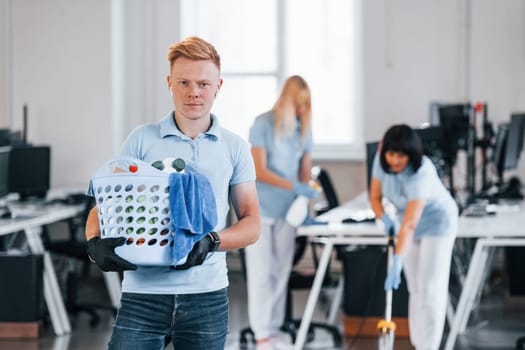 Man holds basket. Group of workers clean modern office together at daytime.