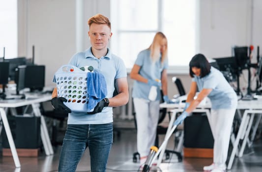 Man holds basket. Group of workers clean modern office together at daytime.