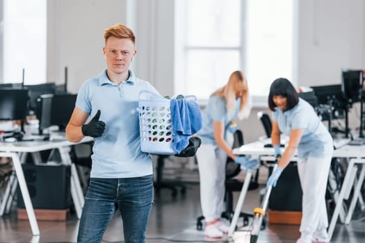 Man holds basket. Group of workers clean modern office together at daytime.