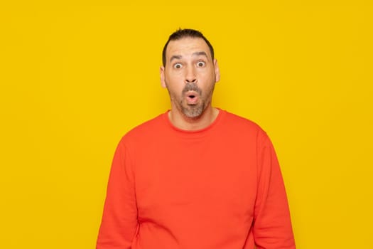 Hispanic man with a beard in a red sweatshirt with his eyes wide with emotion, his mouth is open in surprise generated by something unheard of. Isolated on yellow background
