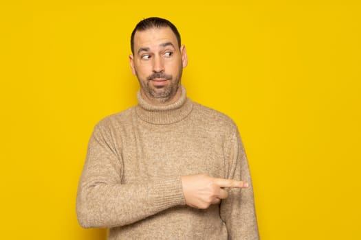 Hispanic man with a beard in a beige turtleneck pointing to the side with his index finger while looking sideways skeptically, he is very curious about what is happening next to him. Isolated