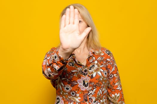 Caucasian blonde woman in a patterned jumper raised her hand in deterrence, campaign to stop violence against women. Woman raised her hand indignantly. Copy space, isolated on yellow background