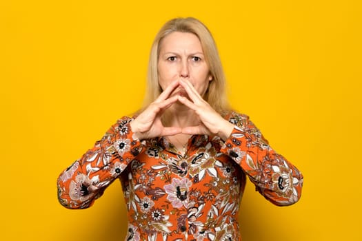 Caucasian blonde woman in a patterned dress clasping her hands in the shape of a triangle in front of her face is hiding an evil and diabolical plan. Isolated on yellow studio background