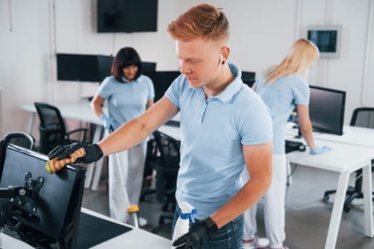 Cleans monitor. Group of workers clean modern office together at daytime.