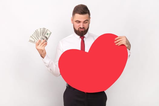 Portrait of young adult bearded man wearing white shirt and tie holding big dollar banknotes, looking at big red heart, choosing money, not love. Indoor shot isolated on gray background.