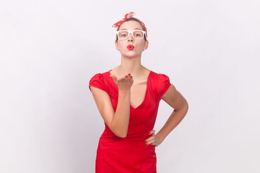 Portrait of pretty lovely cute woman wearing red dress and head band sending air kisses to her boyfriend, keeps han on hip, looking at camera. Indoor studio shot isolated on gray background.
