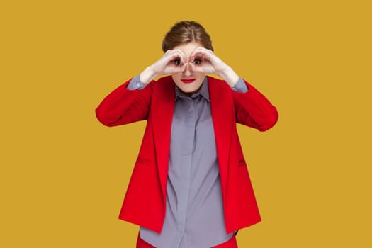 Portrait of amazed concentrated woman with red lips standing making binoculars with her hands, looking far away, wearing red jacket. Indoor studio shot isolated on yellow background.