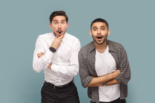 Portrait of two shocked men friends standing together, see something astonished, keeping mouth opened and looking with big eyes. Indoor studio shot isolated on light blue background.