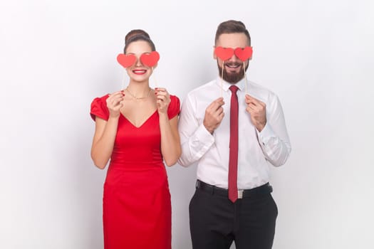 Portrait of romantic man in white shirt and woman in red dress standing together, holding little hearts, covering eye with love symbols on stick. Indoor studio shot isolated on gray background.