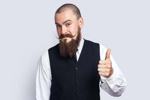 Portrait of pleased friendly man with beard and mustache standing showing thumb up, posing with like gesture, looking at camera. Indoor studio shot isolated on gray background.