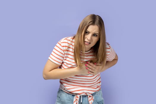 Portrait of blond woman wearing striped T-shirt touching chest, feeling sudden acute pain, risk of heart attack, emergency situation. Indoor studio shot isolated on purple background.
