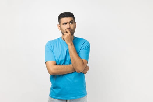 Portrait of serious thoughtful unshaven man wearing blue T- shirt standing keeps hand on his chin, thinking about his work, looking away. Indoor studio shot isolated on gray background.