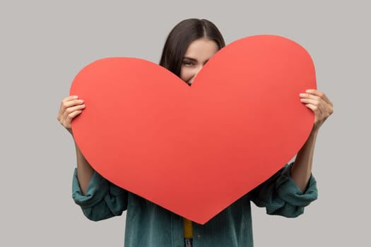 Positive woman with dark hair hiding face behind big red heart and looking at camera, feeling affection fondness, wearing casual style jacket. Indoor studio shot isolated on gray background.
