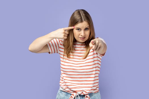 Portrait of displeased blond woman wearing striped T-shirt holding finger near temple and pointing on you, showing stupid gesture, mocking. Indoor studio shot isolated on purple background.