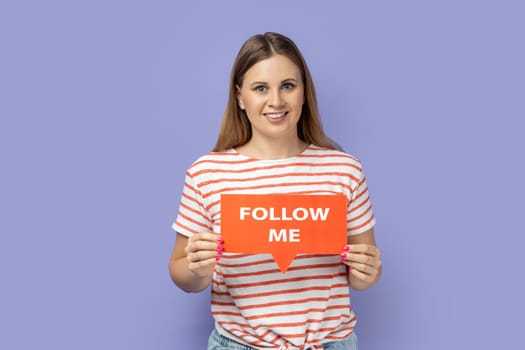 Portrait of pretty cute adorable blond woman blogger wearing striped T-shirt holding red card with follow me inscription, subscribe my vlog. Indoor studio shot isolated on purple background.