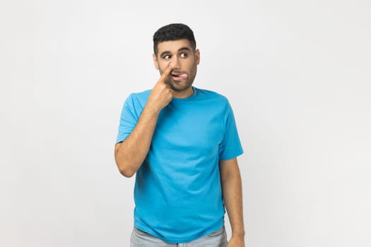 Portrait of funny unshaven man wearing blue T- shirt standing putting finger into his nose, fooling around, bad habits, disrespectful behavior. Indoor studio shot isolated on gray background.