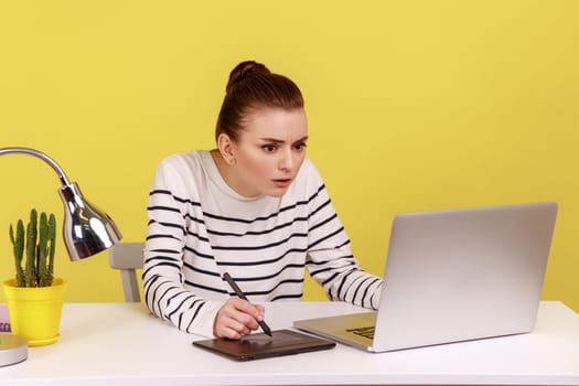 Concentrated busy serious woman working on graphic tablet, drawing sketches, sitting at workplace with laptop looking at display. Indoor studio studio shot isolated on yellow background.