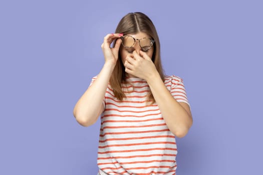 Portrait of frustrated blond woman wearing striped T-shirt touching closed eyes, crying from depression, grief or pain, feeling hopeless. Indoor studio shot isolated on purple background.