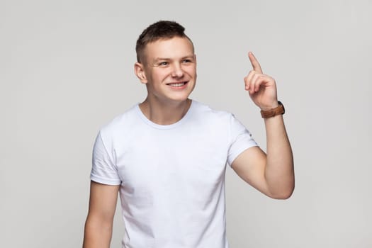 Eureka. Portrait of handsome excited inspired teenager boy wearing T-shirt standing with raised finger, having an excellent idea, smiling. Indoor studio shot isolated on gray background.