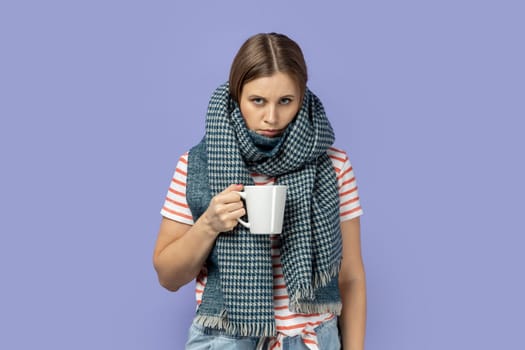 Portrait of sick ill blond woman wearing striped T-shirt and wrapped in warm scarf, posing with sad upset expressing drinking cup of tea. Indoor studio shot isolated on purple background.