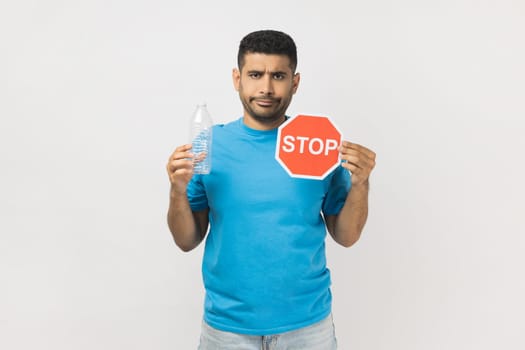 Portrait of sad serious unshaven man wearing blue T- shirt standing holding red stop symbol and empty plastic bottle, Earth protection. Indoor studio shot isolated on gray background.