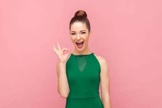 Portrait of excited amazed young adult woman showing ok sign, looking at camera with positive look, expressing happiness, wearing green dress. Indoor studio shot isolated on pink background.