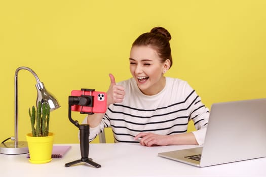 Satisfied joyful woman blogger recording video for followers, looking at phone camera, showing thumb up to her subscribers. Indoor studio studio shot isolated on yellow background.