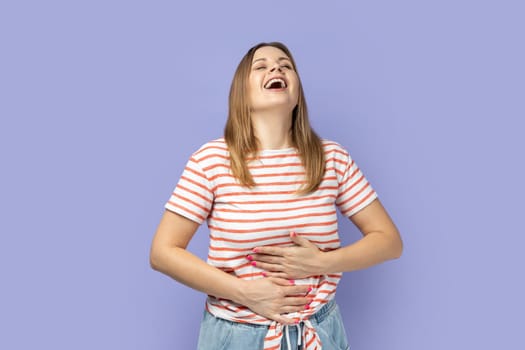 Extremely happy blond woman wearing striped T-shirt holding her stomach and laughing out loud, chuckling giggling at amusing anecdote, sincere emotion. Indoor studio shot isolated on purple background