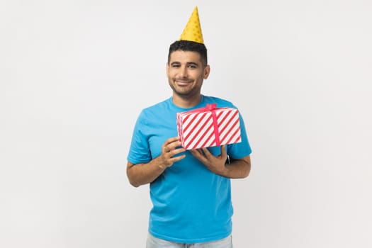 Portrait of festive smiling unshaven man wearing blue T- shirt and yellow party cone standing with present box, celebrating his birthday. Indoor studio shot isolated on gray background.