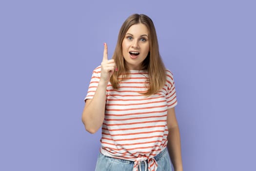 Portrait of inspired blond woman wearing striped T-shirt pointing finger up and looking amazed about sudden genius idea, got solution. Indoor studio shot isolated on purple background.