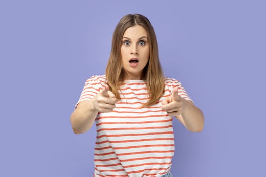 Portrait of blond woman wearing striped T-shirt standing with open mouth pointing to you, making choice and expressing astonishment. Indoor studio shot isolated on purple background.