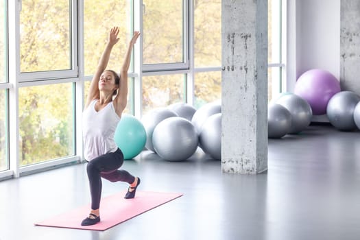 Portrait of sporty flexible woman doing sport lunge exercise, raising hands, keeps eyes closed, being concentrated, warming up training muscle. Indoor shot with window on background.