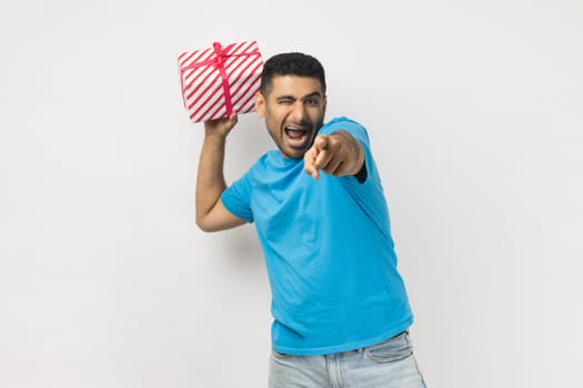 Portrait of excited positive unshaven man wearing blue T- shirt standing throwing present box, pointing finger to camera, gift for you. Indoor studio shot isolated on gray background.