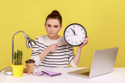 Serious bossy woman office worker pointing finger at big wall clock, time management, looking at camera with strict expression. Indoor studio studio shot isolated on yellow background.