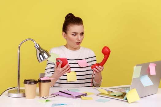 Shocked woman office worker sitting at workplace covered with sticky notes and holding mobile phones in hands and handset. Indoor studio studio shot isolated on yellow background.