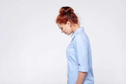 Side view portrait of sad upset redhead woman wearing blue shirt crying with sorrow, feeling lonely hopeless, desperate emotions, worried about troubles. Indoor studio shot isolated on gray background