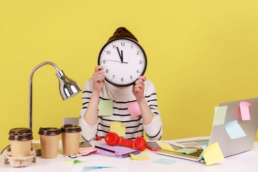 Woman office worker covered with sticky notes sitting at workplace hiding face behind big wall clock, schedule and business meeting appointment. Indoor studio studio shot isolated on yellow background