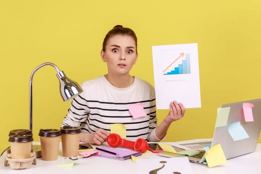 Portrait of serious woman office manager looking at camera showing growth diagram, financial and economic growth of his business. Indoor studio studio shot isolated on yellow background.