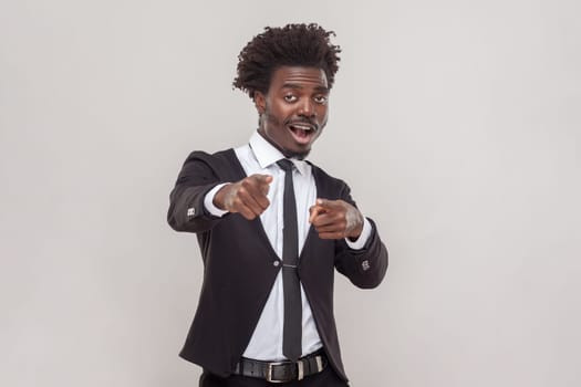 Shocked man with Afro hairstyle pointing index finger at camera with great surprise, holds breath indicates forward, wearing white shirt and tuxedo. Indoor studio shot isolated on gray background.