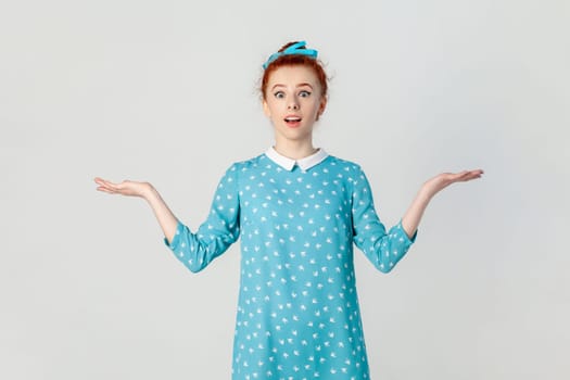 Portrait of shocked confused ginger woman standing with spread hands, looking at camera with big eyes, dosen't know how to react, wearing blue dress. Indoor studio shot isolated on gray background.