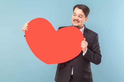 Portrait of satisfied cheerful man standing showing big read heart, confesses his feelings to someone, wearing black suit with red tie. Indoor studio shot isolated on light blue background.