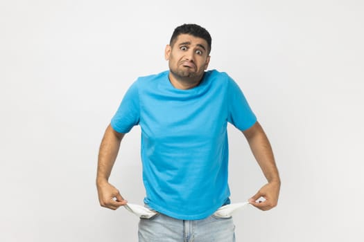 Portrait of sad poor attractive unshaven man wearing blue T- shirt standing showing his empty pockets, has no money, lost his job. Indoor studio shot isolated on gray background.