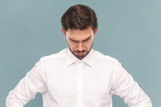 Portrait of pensive strict bully man with mustache with hands on hips, looking from under the forehead, being angry, wearing Indoor studio shot isolated on light blue background.