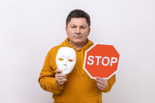 Portrait of serious dark haired man holding white mask with unknown face and red traffic sign, looking at camera, wearing urban style hoodie. Indoor studio shot isolated on white background.