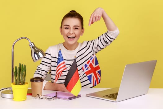 Excited happy woman pointing at flags of usa, german and great britain, bragging with knowing of many languages, polyglot. Indoor studio studio shot isolated on yellow background.